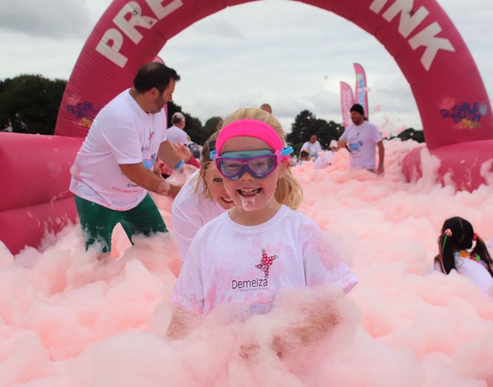 A young girl is standing in a sea of pink bubbles, wearing a pink headband and swimming goggles.