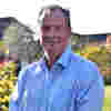Chair Sean Reynolds CB CBE DFC stands smiling in sunny gardens, wearing a long-sleeved pale blue shirt.