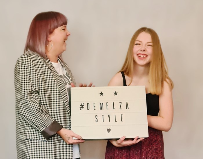 Two girls are smiling and laughing, hold a sign which reads 'Demelza Style'.