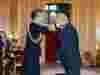 Person in navy uniform places medal on man in suit during formal ceremony in ornate room with gold decor and red and gold chairs.