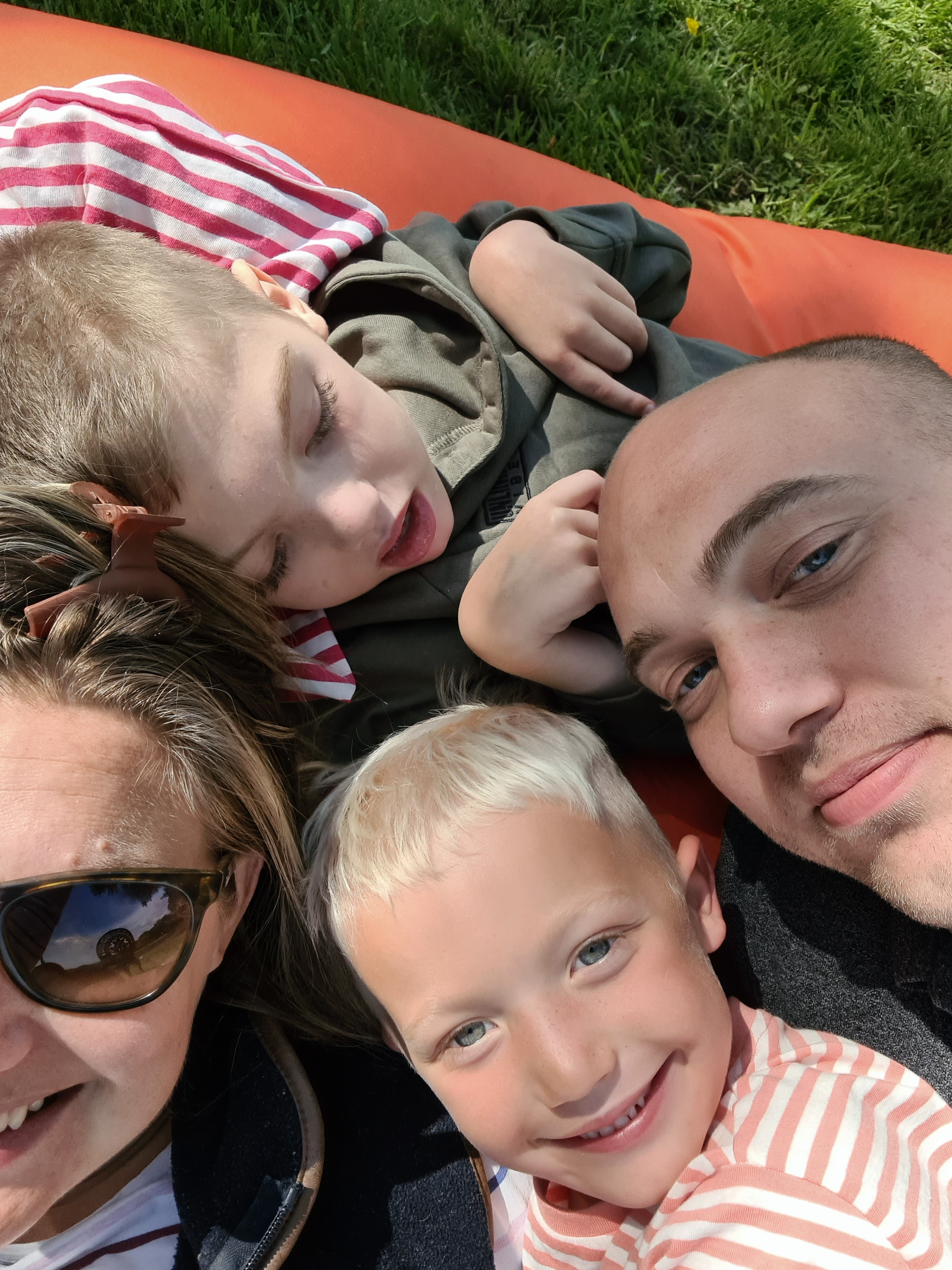Two adults and two children lying on an orange mat outdoors, close-up of faces and upper bodies; some wear striped clothing, with green grass in the background.