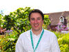 Man in white shirt and teal lanyard standing in front of a garden with green foliage and purple flowers.