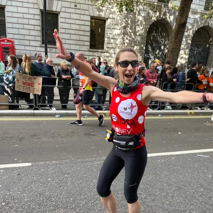 A woman wearing a Demelza running vest, holds her hands in the air, smiling as she runs in the London Marathon.