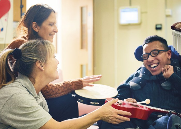 A demelza Health Care Assistant, with a parent and a teenager in his mobility scooter, playing with a ukulele while laughing 