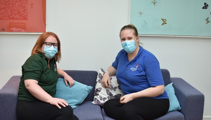 A registered nurse and a health care assistant sit on a grey sofa, wearing medical masks.