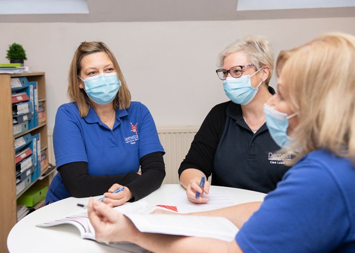 Three nurses, wearing blue polo tops and face coverings, sit around a round table, discussing paperwork.