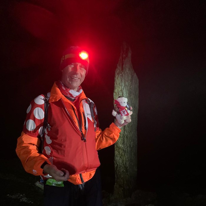 A man wearing a orange waterproof jacket and a head torch, at the summit of Mount Snowdon.