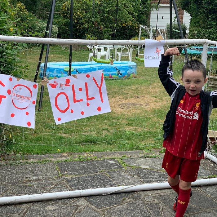 A boy celebrates completing his fundraising challenge. He is standing in front of a football goal with a 'GO OLLY' sign.
